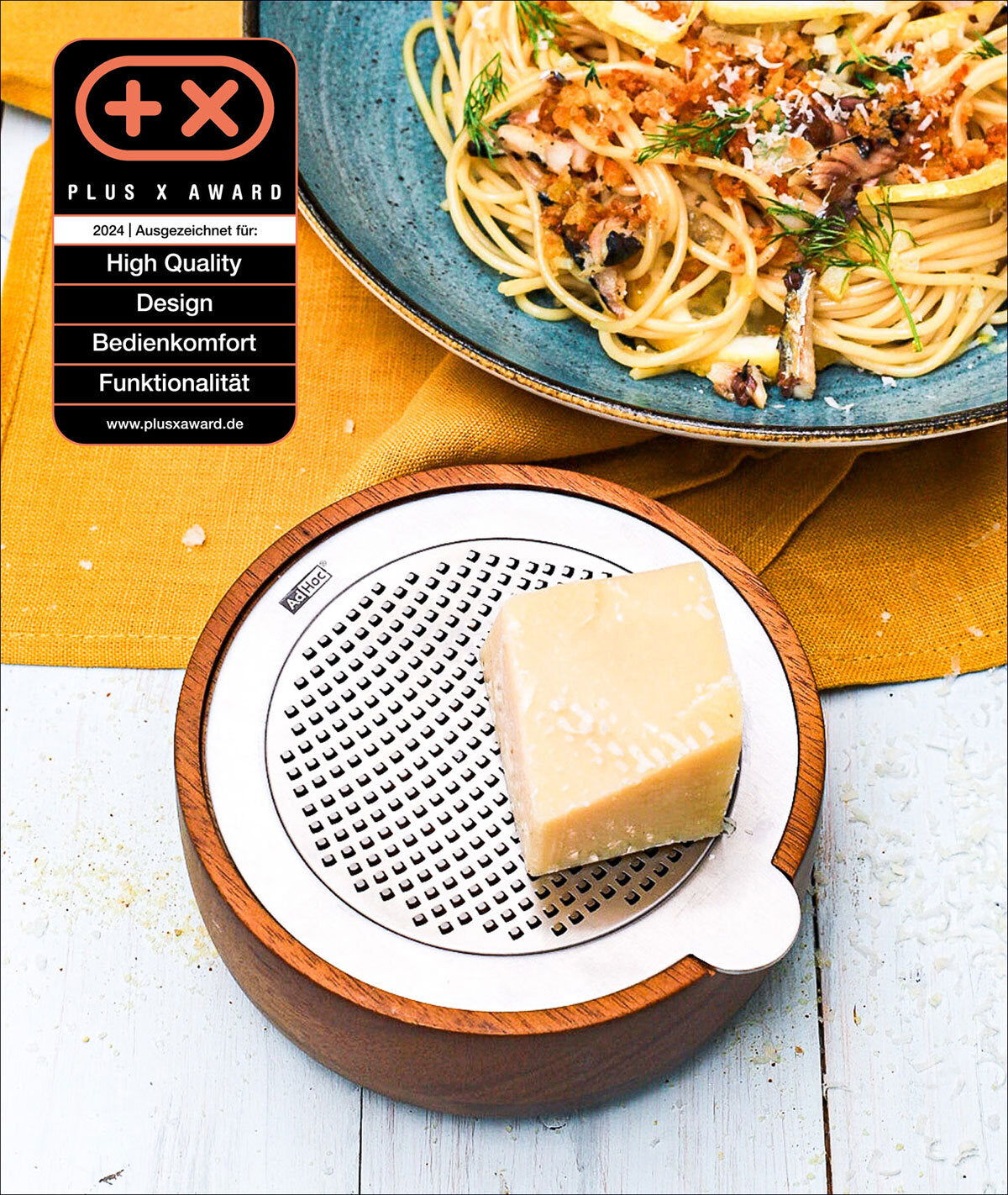 Käsestück auf einer runden, weißen Käsehobel mit Holzrahmen, daneben Pasta mit Kräutern auf blauer Platte, auf gelbem Tuch.
