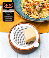 Käsestück auf einer runden, weißen Käsehobel mit Holzrahmen, daneben Pasta mit Kräutern auf blauer Platte, auf gelbem Tuch.