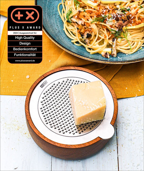 Käsestück auf einer runden, weißen Käsehobel mit Holzrahmen, daneben Pasta mit Kräutern auf blauer Platte, auf gelbem Tuch.