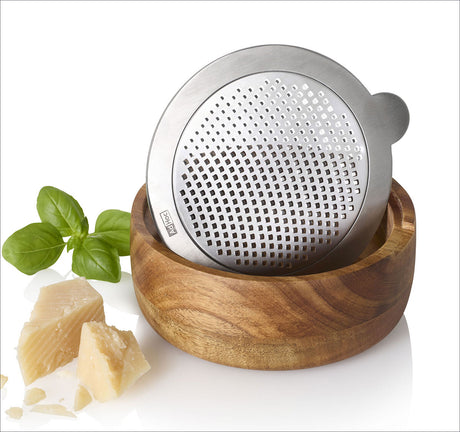 Rostfreier Käsehobel in Holzschale, daneben Basilikum und Parmesan.