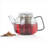 Glass-Kanne mit rotem Tee, Teesieb im Inneren, daneben lose Teeblätter auf weißem Hintergrund.