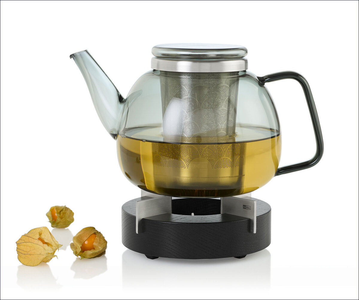 Glas-Teekanne mit heißem Tee, Teesieb im Inneren, auf Heizplatte, daneben Physalis-Früchte.