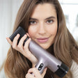 Junge Frau mit braunen Haaren hält einen Haartrockner an ihr Ohr, lächelt und schaut in die Kamera.