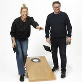 Zwei lachende Personen spielen Cornhole, eine Frau wirft eine Tasche, beide tragen schwarze Kleidung, im Hintergrund weiß.