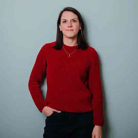 Frau mit dunklen Haaren, rotem Pullover, schwarzer Hose, steht vor hellblauer Wand, schaut nach rechts.