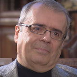 Ein Mann mit Brille, grauem Haar, trägt einen schwarzen Rollkragen und eine graue Jacke.