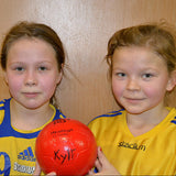 Zwei Mädchen in blauen und gelben Trikots halten einen roten Handball.