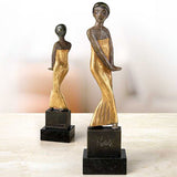 Zwei Skulpturen von Frauen in goldenen Kleidern auf schwarzen Basen, eine größere und eine kleinere Figur.