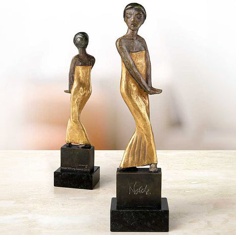 Zwei Skulpturen von Frauen in goldenen Kleidern auf schwarzen Basen, eine größere und eine kleinere Figur.