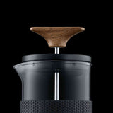 Kaffeemühle mit Holzknopf, transparentem Glasbehälter und schwarzem Griff vor schwarzem Hintergrund.