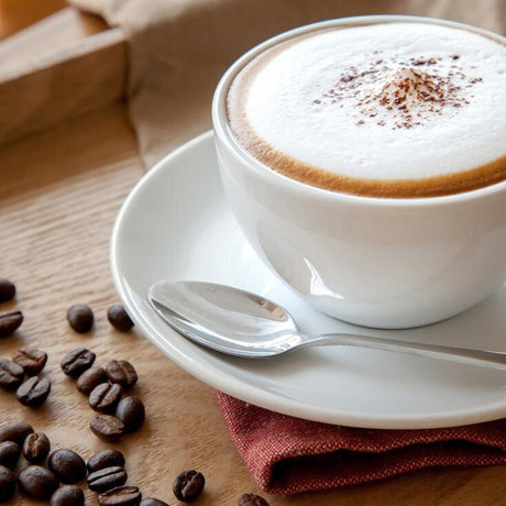 Weiße Kaffeetasse mit Cappuccino, Schaum und Kakaopulver, auf weißem Teller mit Löffel, umgeben von Kaffeebohnen auf Holz.