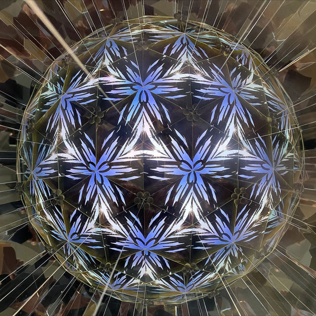 Blick nach oben auf eine geometrische, leuchtende Kugel mit blauen und weißen Mustern, umgeben von reflektierenden Flächen.