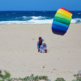 Ein Kind sitzt mit einem Erwachsenen am Strand, im Hintergrund das Meer, und ein farbenfroher, aufgespannter Kite.