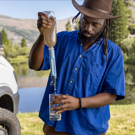 Mann mit Hut füllt Wasser aus einer Flasche mit Wasserfilter in ein Glas im Freien.