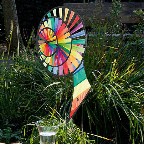 Buntes Windspiel mit Spiral-Design und Farbsegmenten, im Garten neben Pflanzen und einem Glas Wasser.