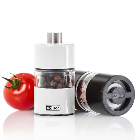 Zwei Salz- und Pfeffermühlen, eine Tomate und Salz- und Pfefferkörner auf weißem Hintergrund.
