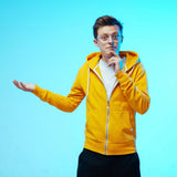 Junger Mann in gelber Kapuzenjacke, trägt Brille, steht vor blauem Hintergrund, wirkt nachdenklich, mit einer Hand gestikuliert.
