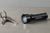 Schwarze Zweibrüder 1T Mini-Taschenlampe mit runder Linse neben zwei Schlüsseln auf einer grauen Fläche