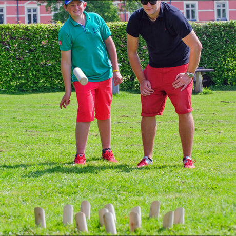Zwei Personen spielen Kubb im Garten, eine Person wirft einen Holzblock, die andere beobachtet.