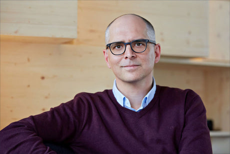 Mann mit Brille, blauer Hemd und dunkelrotem Pullover vor Holzregal.