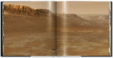 Dünes, trockene Wüstenlandschaft mit Felsen im Hintergrund, offene, sandige Fläche im Vordergrund.